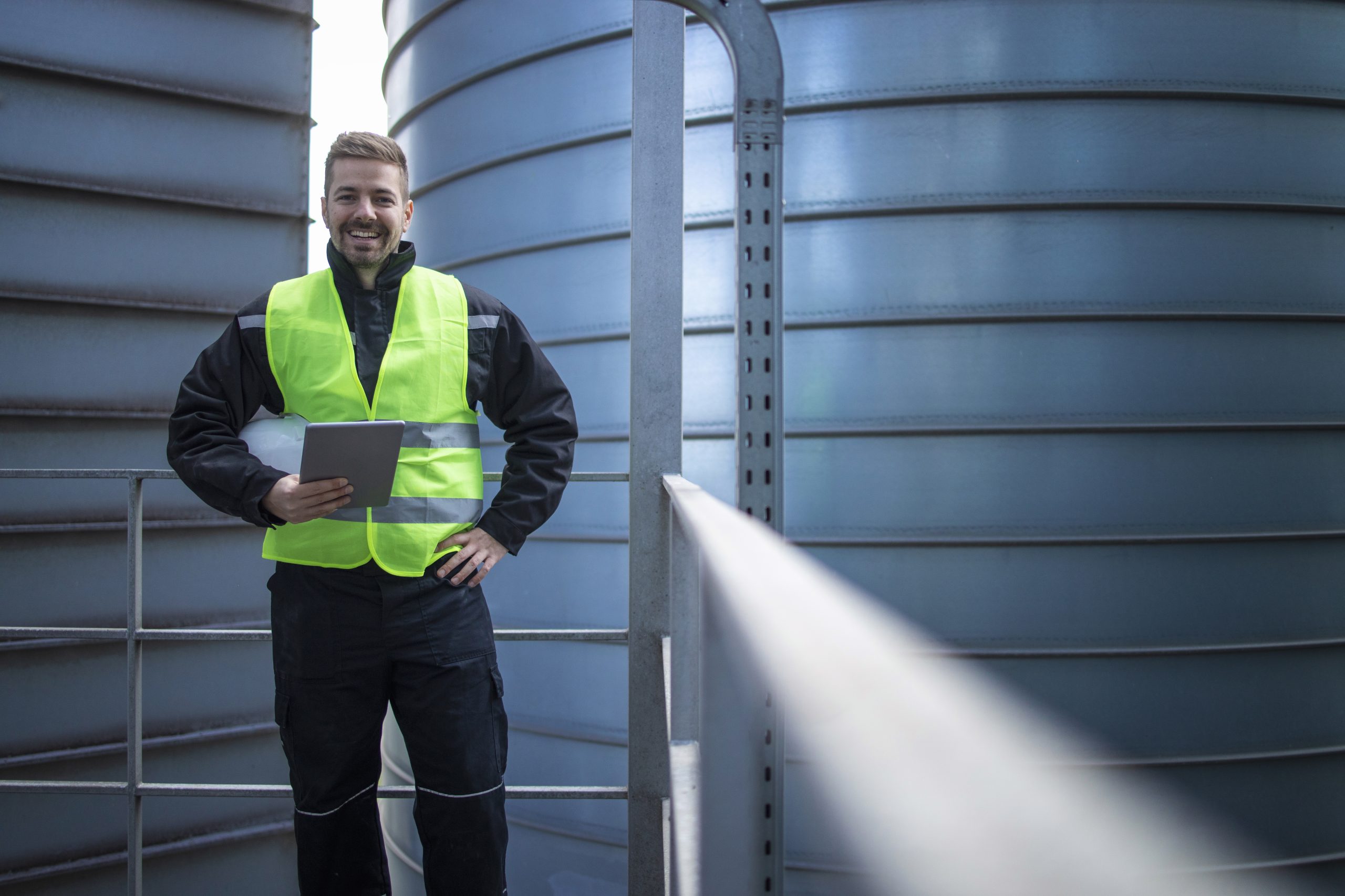 Portrait of factory engineer worker standing on metal platform between industrial storage tanks and looking to the camera.